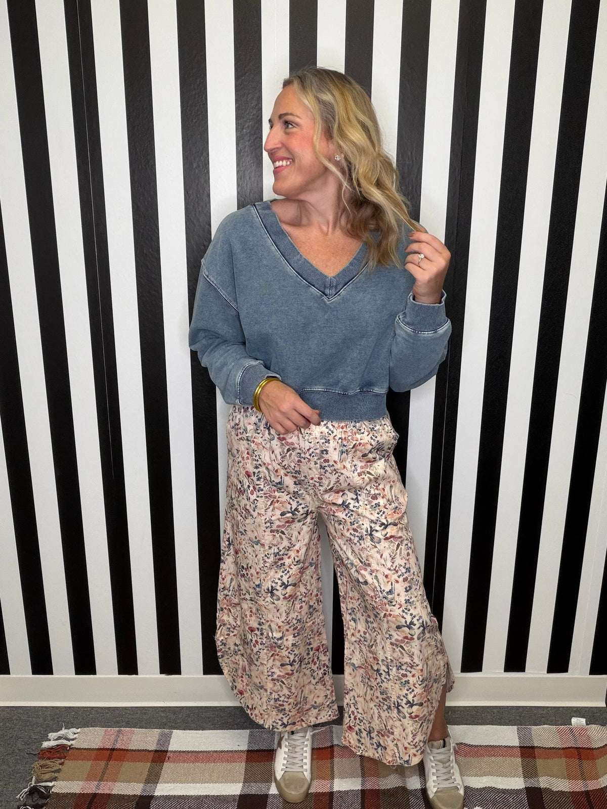 A woman stands in front of a black and white striped wall wearing a washed blue v-neck long-sleeve top, wide-leg floral pants, and white sneakers.