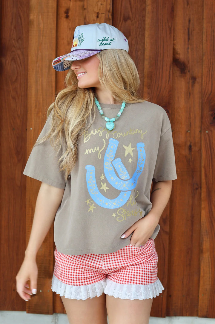Woman wearing a brown t-shirt with a graphic design, red and white checkered shorts, and a cap against a wooden background.