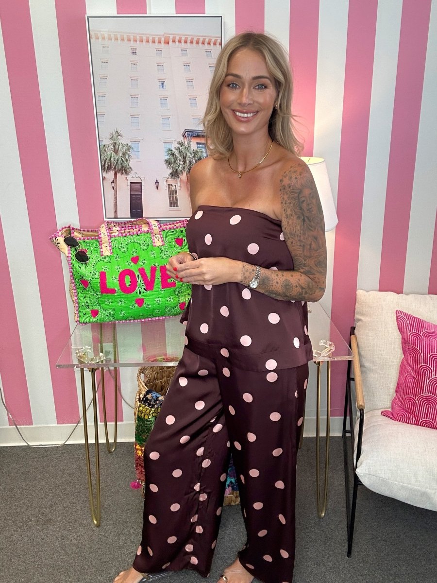 A woman wearing a dark brown strapless jumpsuit with light pink polka dots stands in a room with pink and white striped walls. She is holding a bright green handbag with pink 'LOVE' lettering.