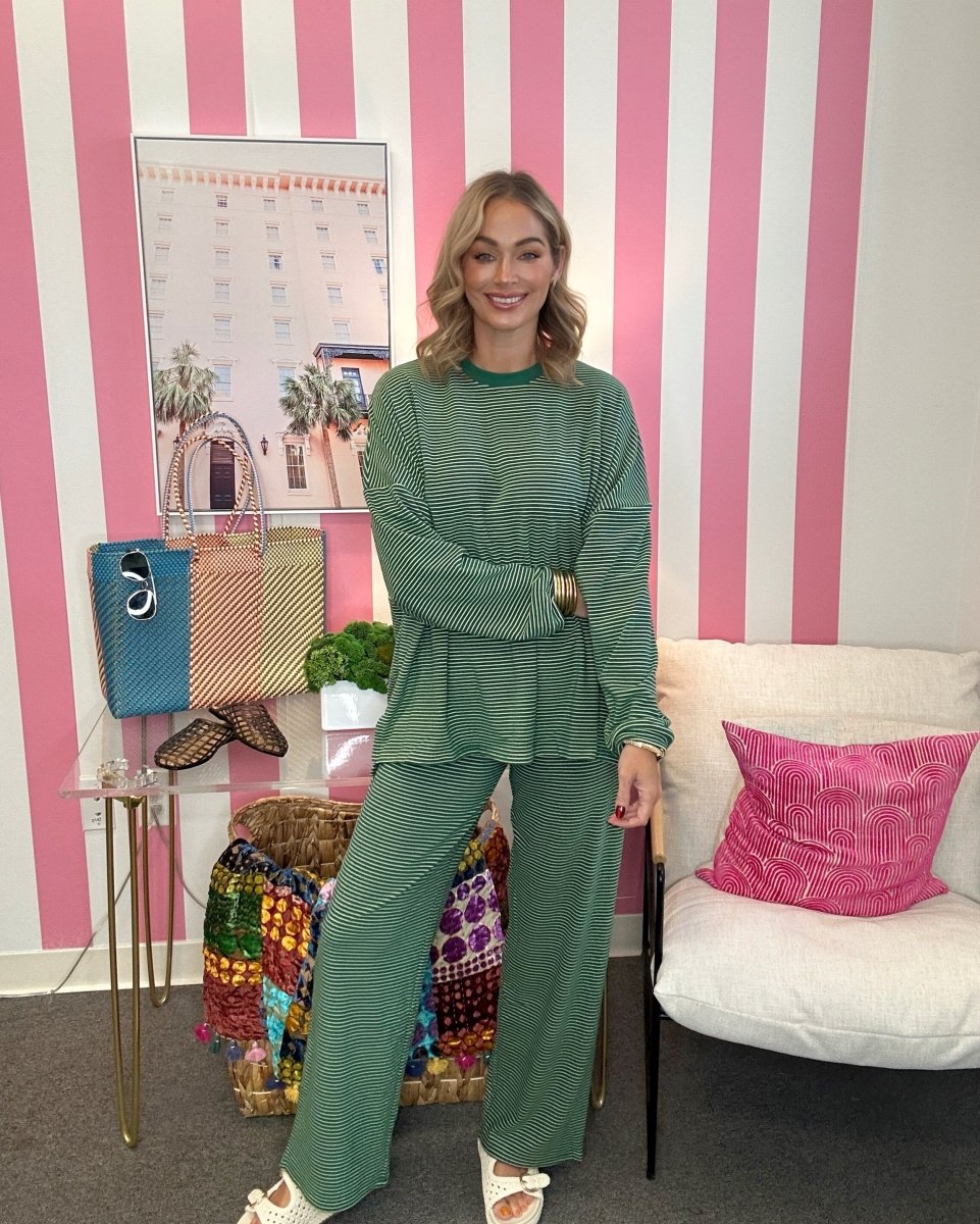 A woman modeling a matching sage green ribbed knit lounge set consisting of a long-sleeve top and wide-leg pants, standing in a room with pink and white striped walls.