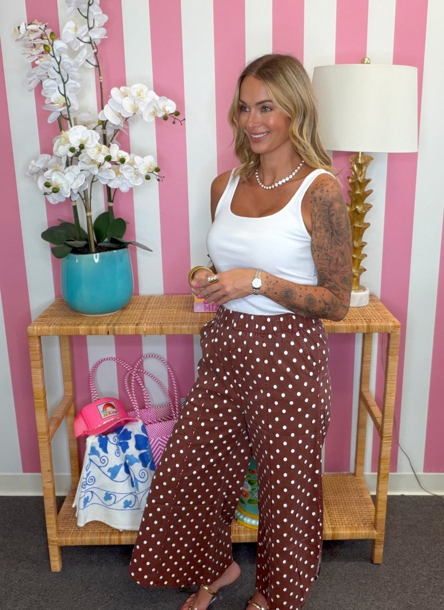 A woman stands indoors in front of a pink and white striped wall, wearing a white tank top and brown wide-leg pants with white polka dots. She is posed beside a wicker console table with a turquoise orchid pot, a lamp, and handbags on the lower shelf.