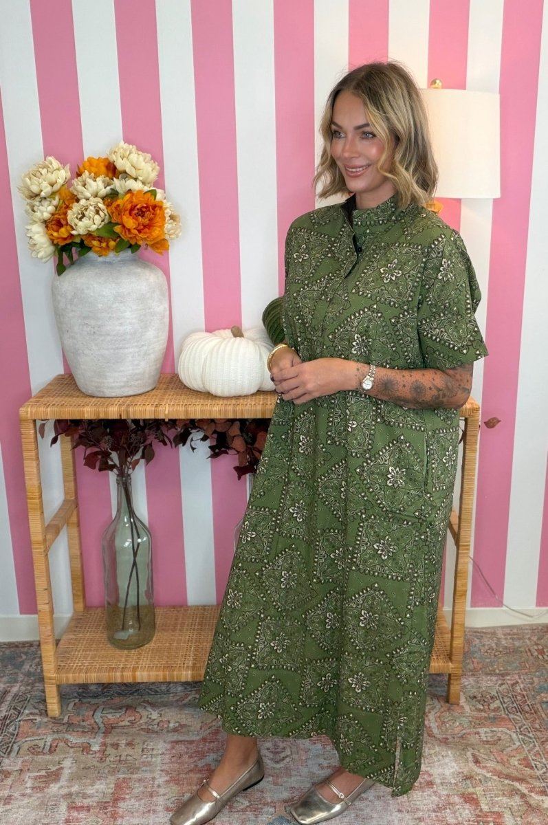 A woman modeling a green embroidered midi dress in front of a pink and white striped wall, standing next to a wooden side table decorated with a vase of orange and white flowers, a white pumpkin, and a glass bottle.