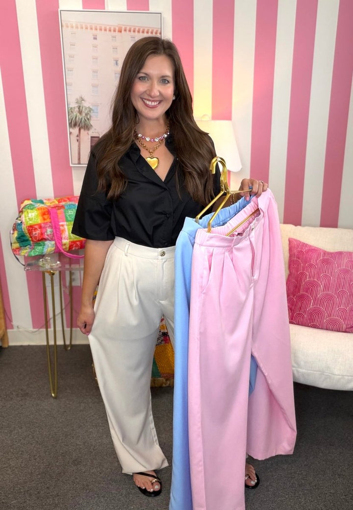 Woman holding pastel-colored pants in a room with striped walls and a colorful bag.
