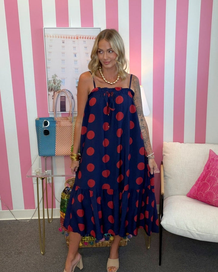 A woman models a navy blue maxi dress with red polka dots, standing in front of a pink and white striped wall. She is holding a colorful patterned clutch bag, with a side table, framed art, and a white sofa visible in the background.