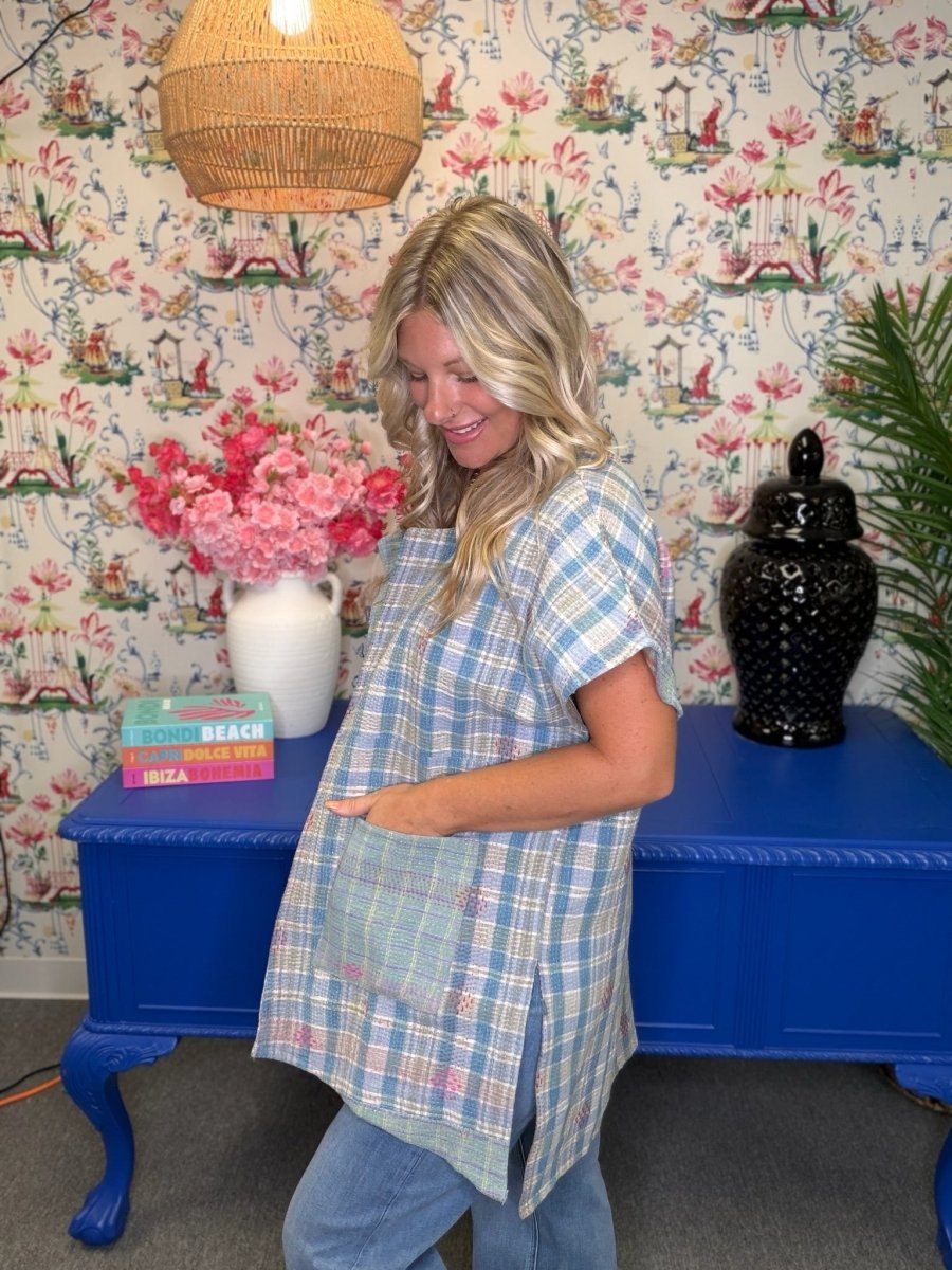 A pregnant woman wearing a light blue and white plaid short-sleeve maternity tunic top, standing in front of a blue console table decorated with pink flowers and a black jar, against a floral wallpaper background.
