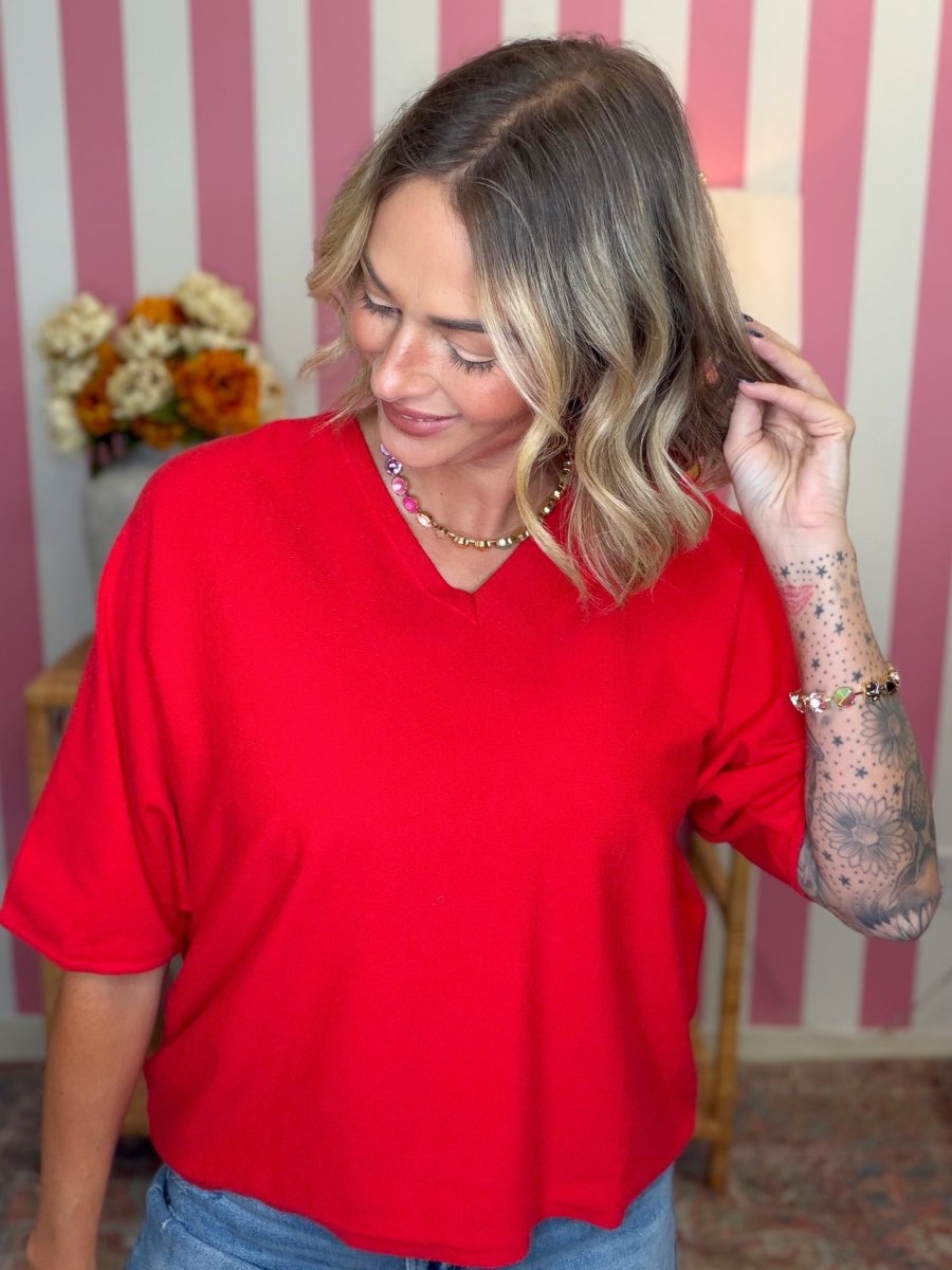 A woman wearing a bright red short-sleeve V-neck top, standing in front of a pink and white striped wall.