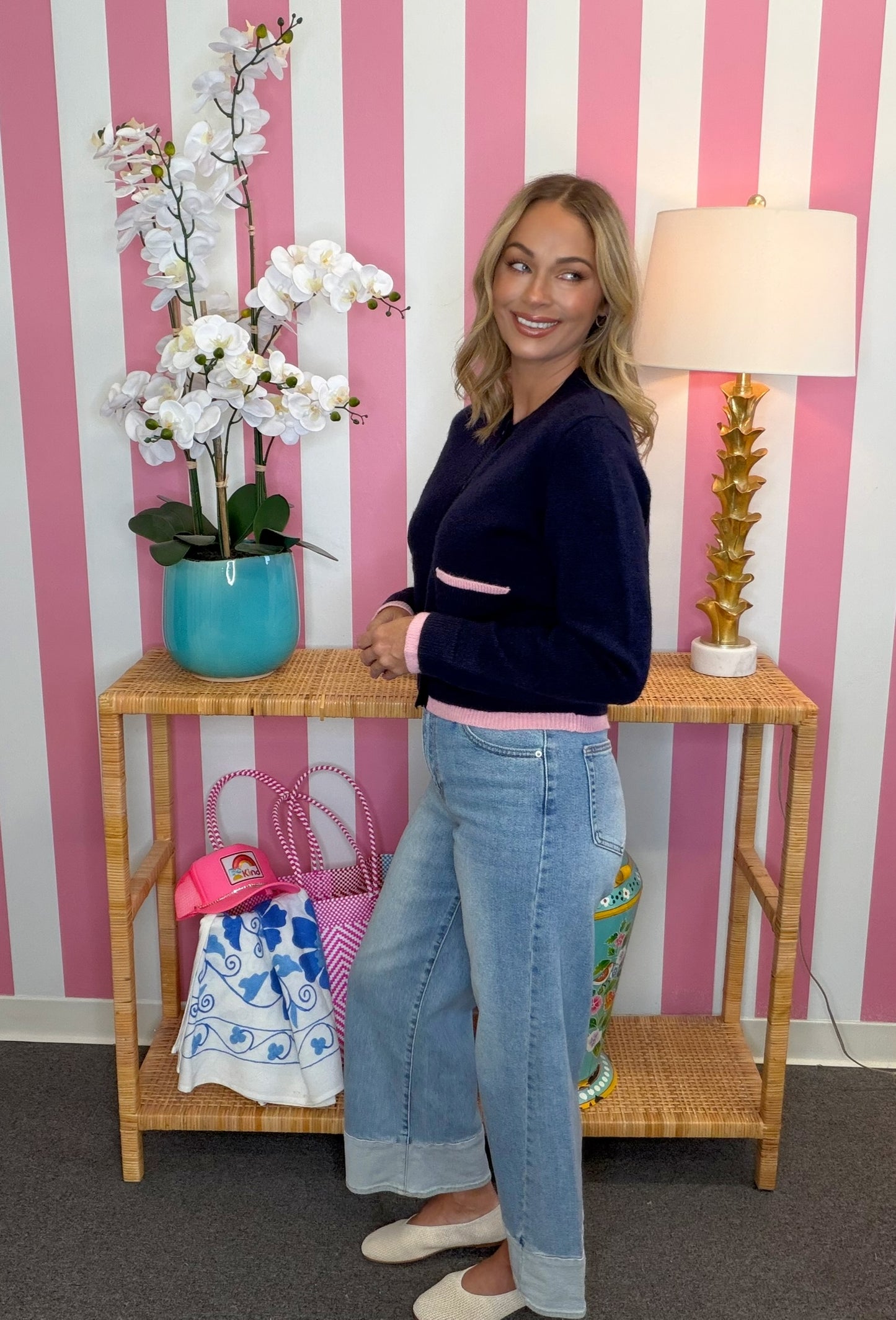 A woman stands in a pink and white striped room wearing a navy blue cropped sweater with pink trim, light blue wide-leg jeans, and white flats. She is posed next to a wooden console table decorated with a potted white orchid, a pink patterned tote bag, and a gold lamp.
