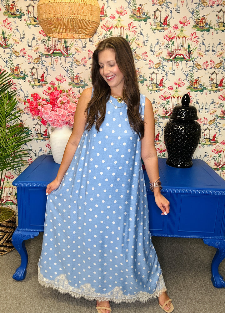 Woman in a blue polka dot dress standing in a room with floral wallpaper and a blue bench.