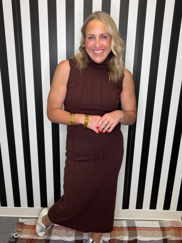 A woman wearing a sleeveless, ribbed brown mock-neck midi dress, standing in front of a black and white striped wall.