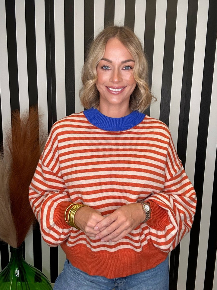 A smiling blonde woman wearing a long-sleeve knit sweater with horizontal orange and white stripes and a solid blue crew neck, standing in front of a black and white striped wall.