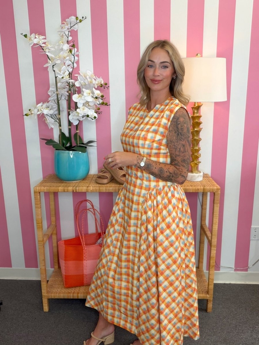 Woman in a checkered dress standing in front of a pink and white striped wall with a table and decorative items.