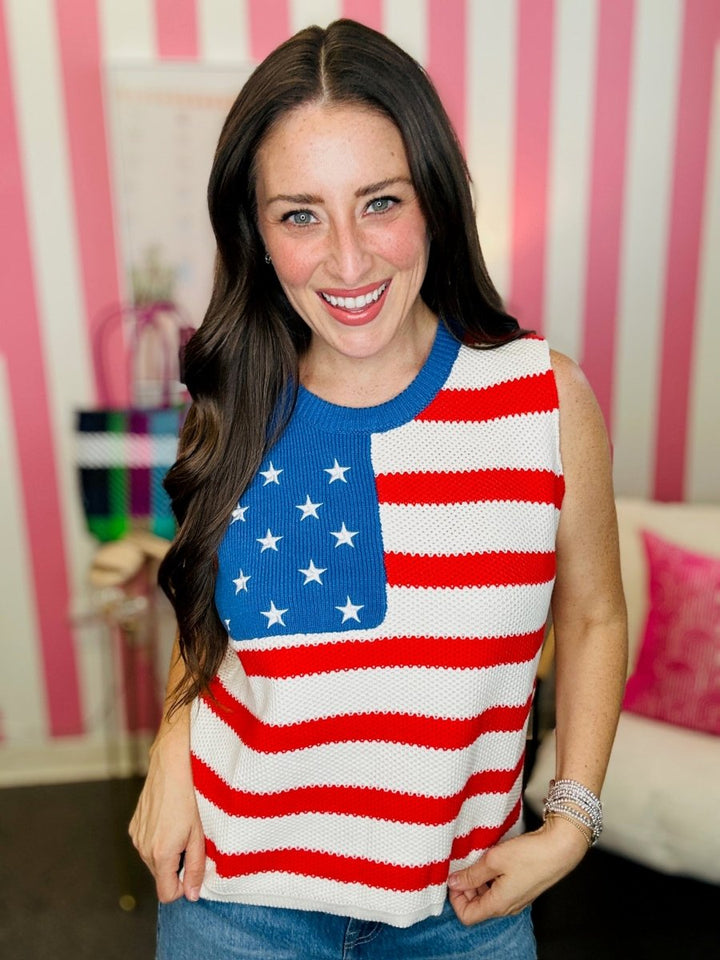Woman wearing a sleeveless sweater with an American flag design in a room with pink and white striped walls.