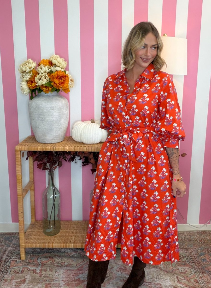 A woman wearing a red midi shirt dress with a white and purple floral print, standing in front of a pink and white striped wall next to a wooden side table with a vase of flowers.