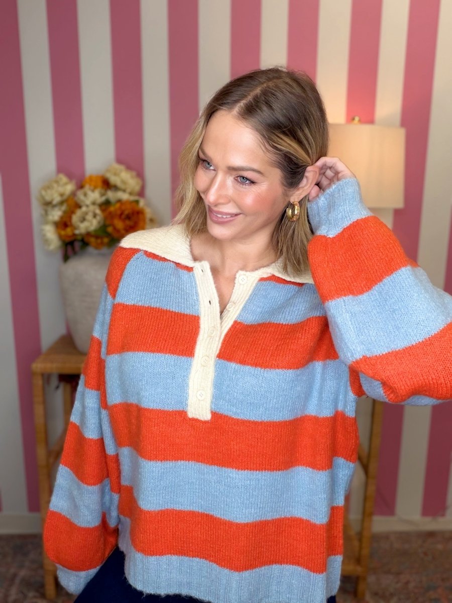 A woman wearing a fuzzy knit long-sleeve henley sweater with horizontal stripes in orange-red and light blue, featuring a cream ribbed collar and placket. She is posing in front of a pink and white striped wall.