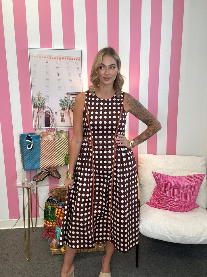 A woman models a brown sleeveless midi dress with white polka dots and orange trim, standing in front of a pink and white striped wall. She is posing with handbags on a side table and a white armchair with a pink pillow in the background.