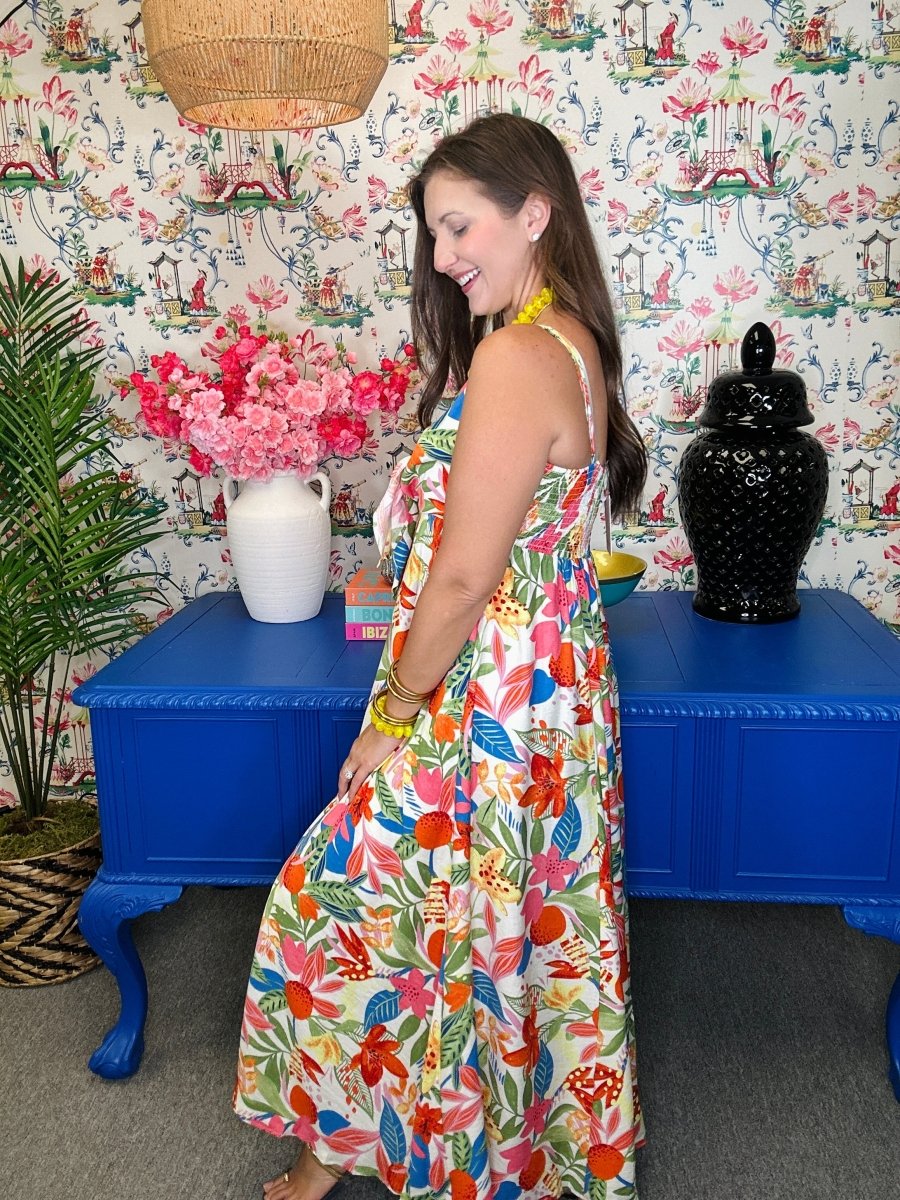 Woman in a colorful floral dress standing in a room with patterned wallpaper and blue tables.