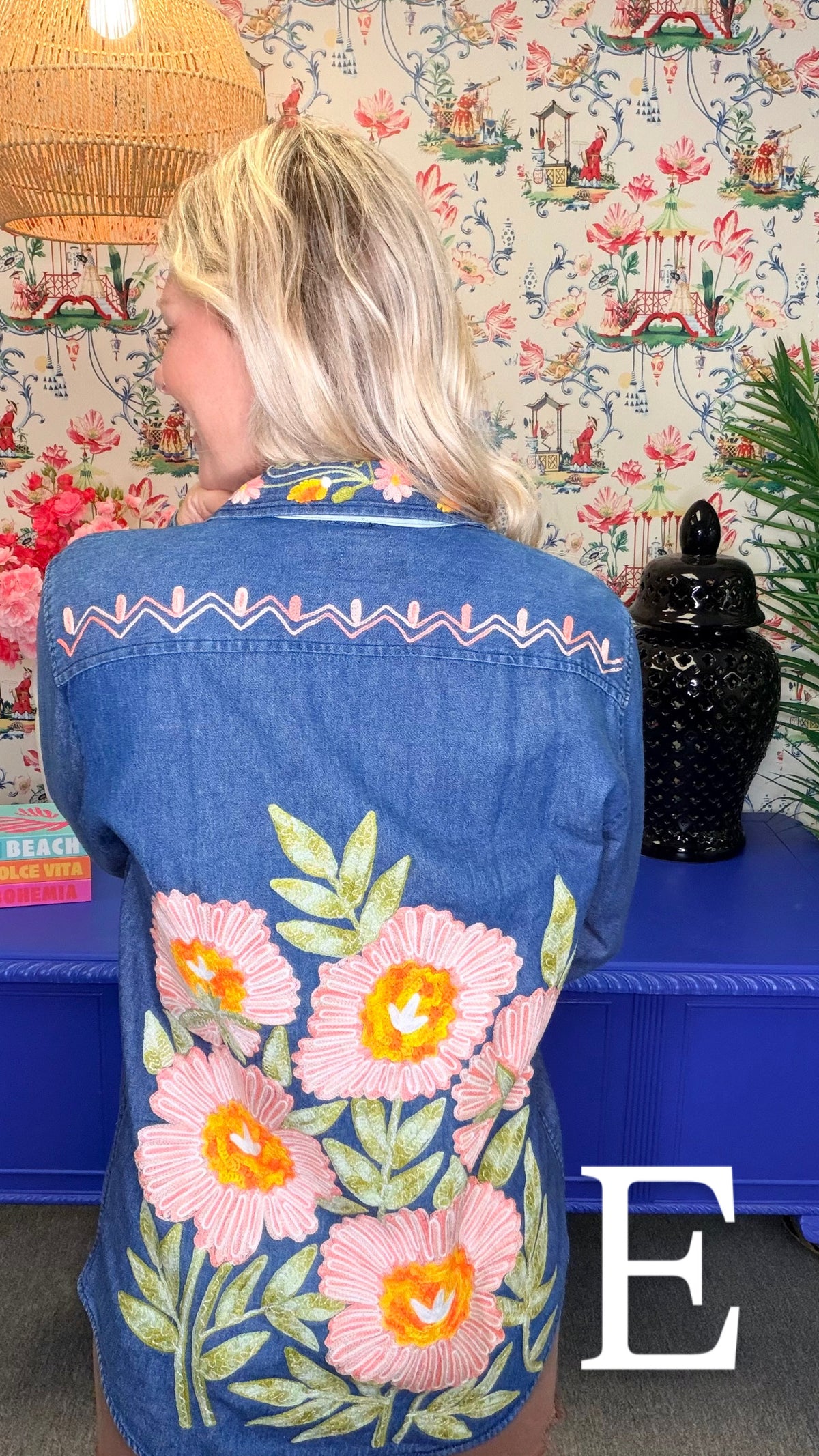 A woman viewed from behind wearing a blue denim shirt jacket with large pink and yellow floral embroidery and green leaves on the back, set against a patterned wallpaper background.
