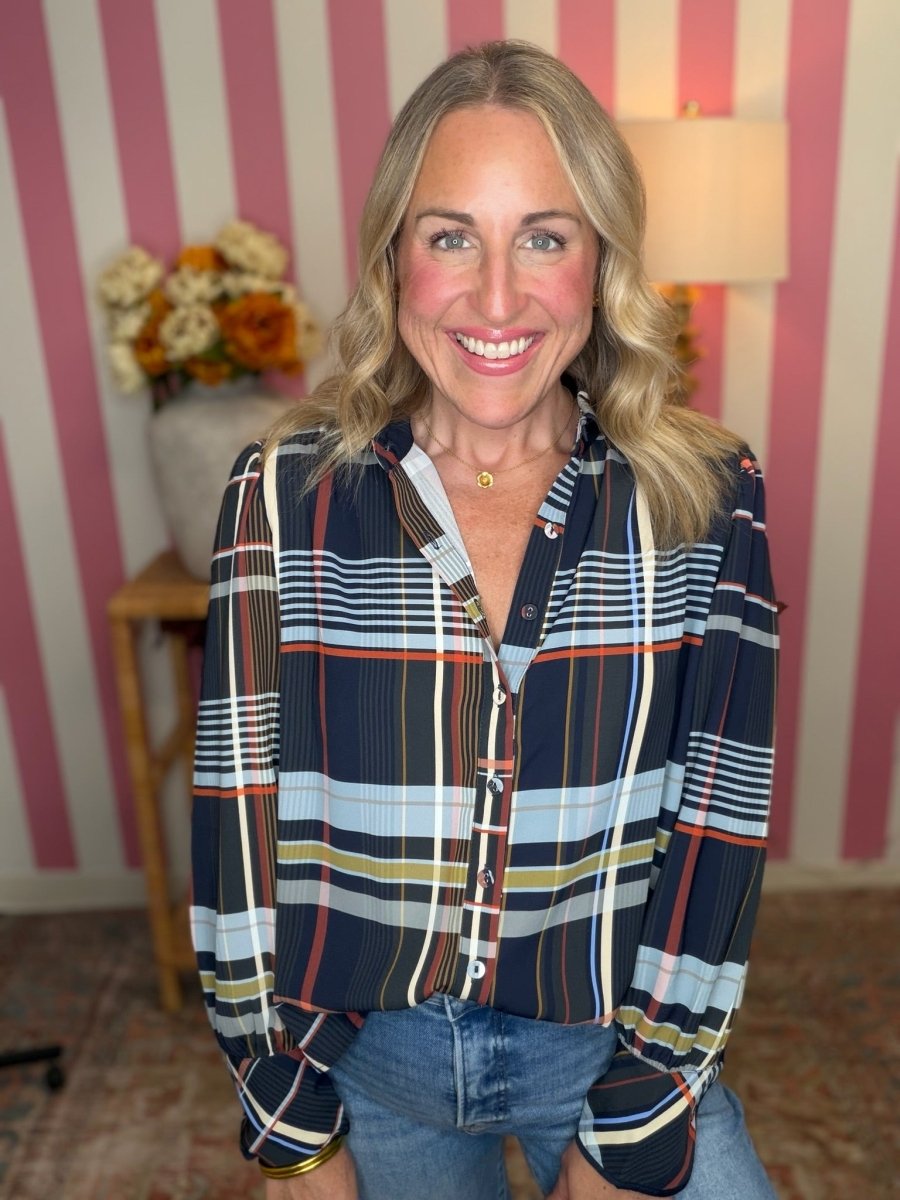 A smiling blonde woman wearing a long-sleeve button-up plaid shirt with navy, light blue, white, orange, and yellow stripes, paired with blue jeans, standing in front of pink and white striped wallpaper.