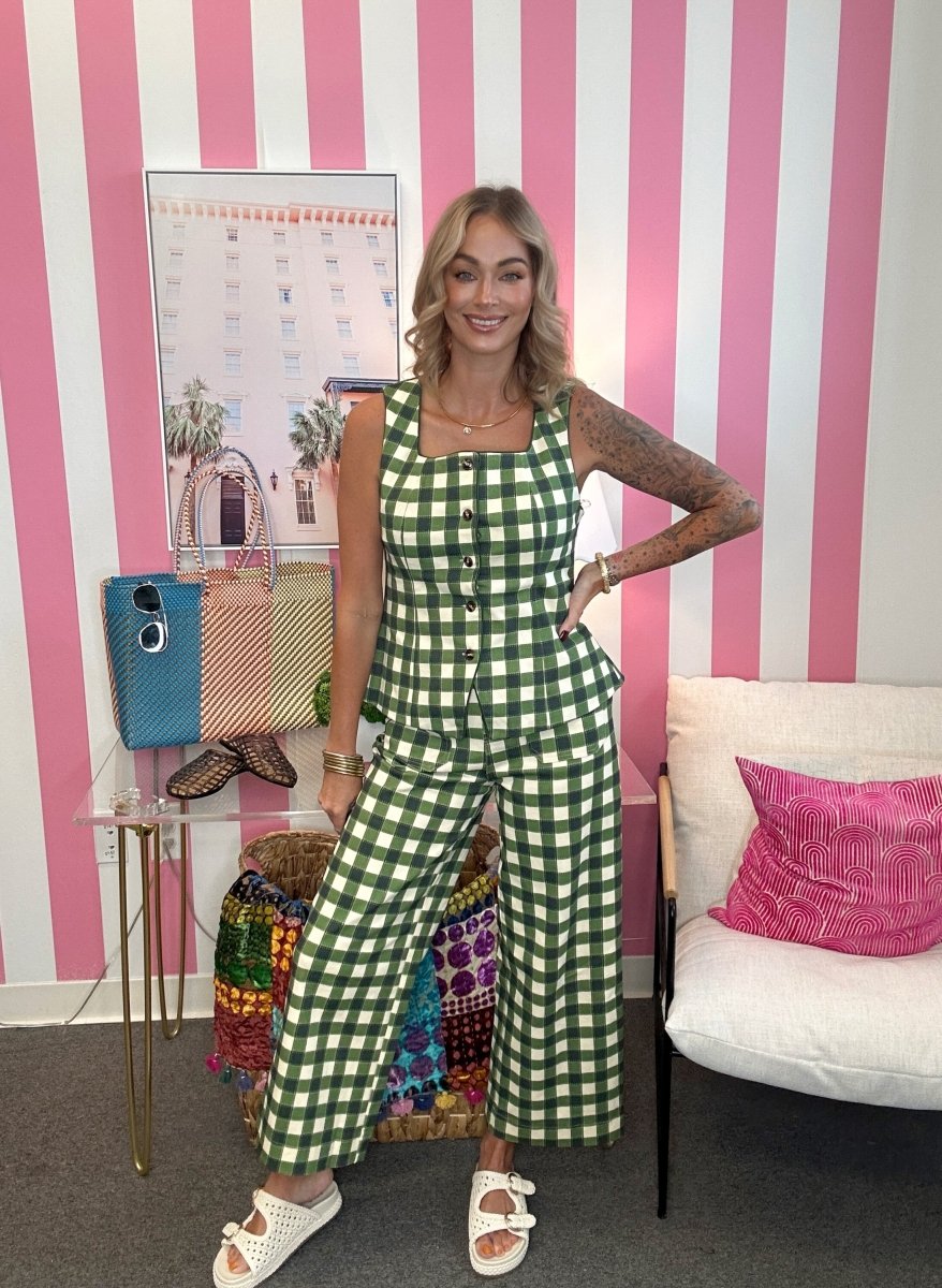 A woman stands in a room with pink and white striped walls wearing a green and white gingham sleeveless jumpsuit. She is posing with her hands on her hips, with a small table, colorful bags, and a white chair with a pink pillow in the background.