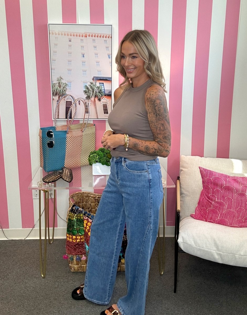 A woman with blonde hair and tattoos stands in a room with pink and white striped walls, wearing a sleeveless taupe top and high-waisted wide-leg blue jeans. She is posing next to a glass table with handbags and a white armchair with a pink pillow.