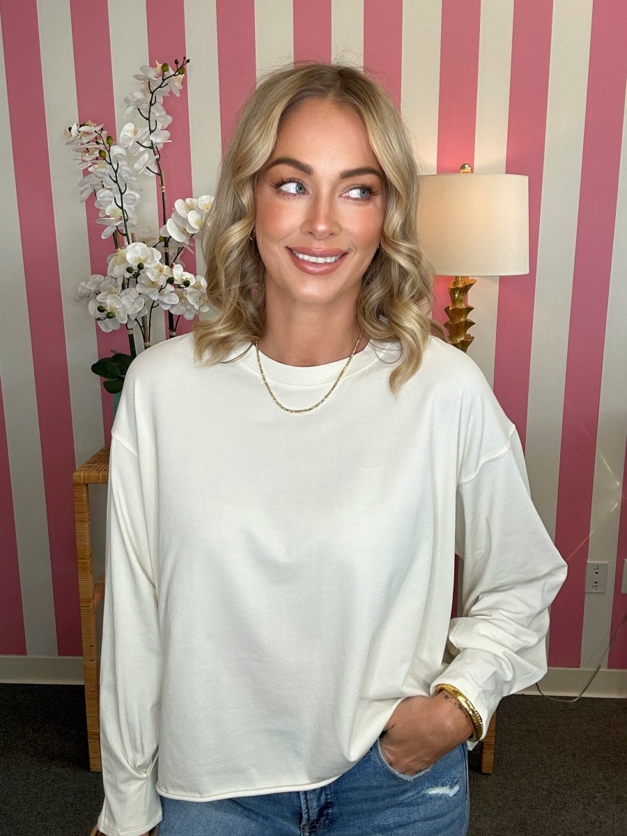 A woman with blonde wavy hair wearing a cream-colored long-sleeve crewneck sweatshirt and blue jeans, standing in front of a pink and white striped wall with a lamp and flowers in the background.