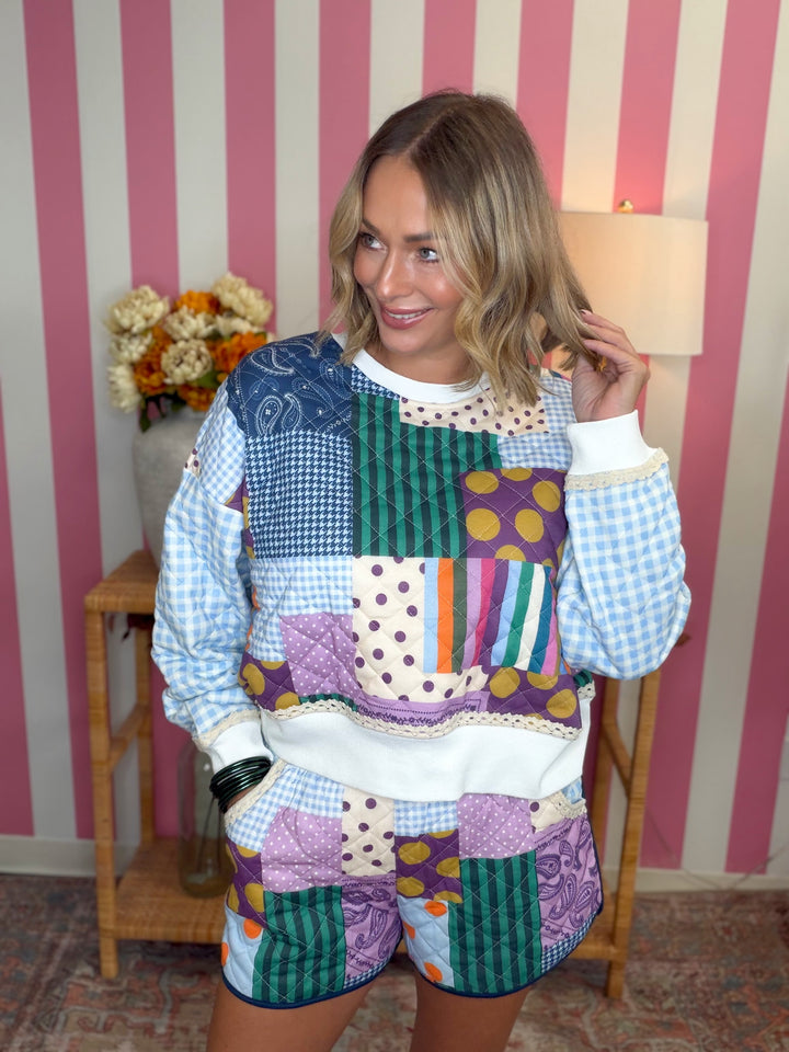 A woman modeling a matching patchwork quilted sweatshirt and shorts set in a room with pink and white striped wallpaper. The set features various colorful patchwork panels including gingham, polka dots, stripes, and quilted fabrics in blue, green, purple, orange, and white.