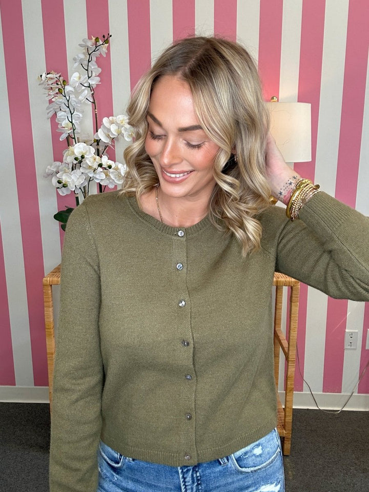 A woman wearing an olive green knit cardigan sweater with silver buttons, paired with blue jeans, standing in front of a pink and white striped wall.