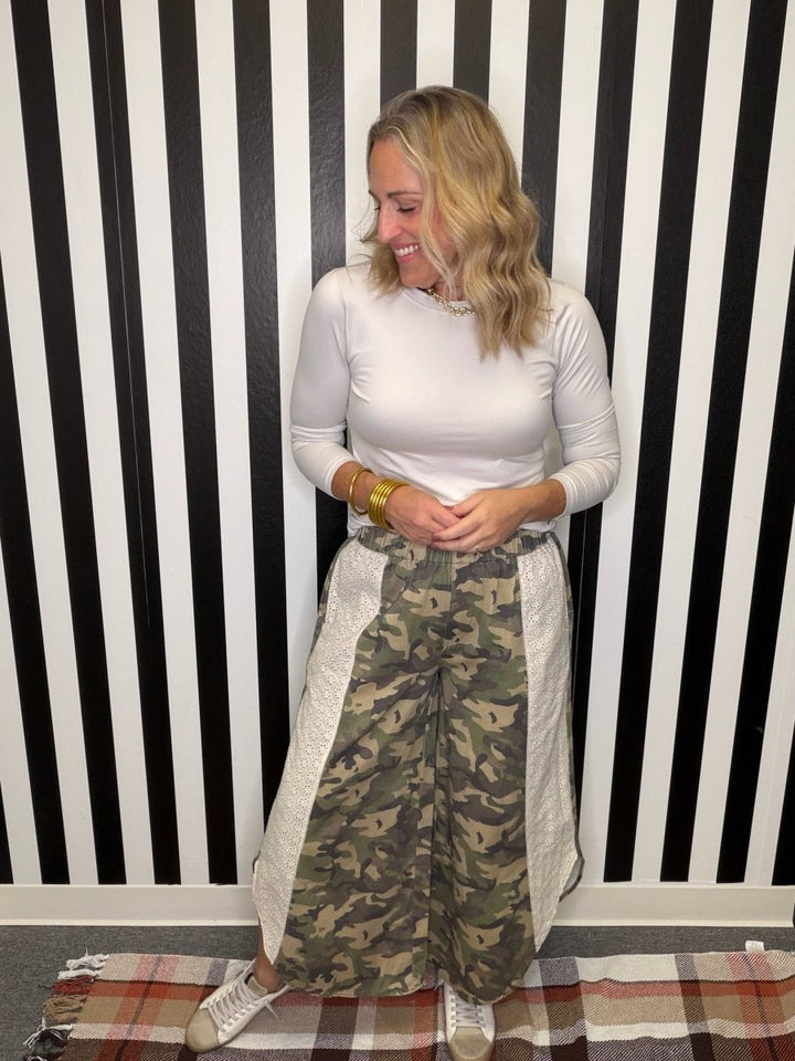 A woman stands in front of a black and white striped wall wearing a white ribbed long-sleeve mock neck top, a long camouflage print skirt with quilted side panels, and white sneakers.