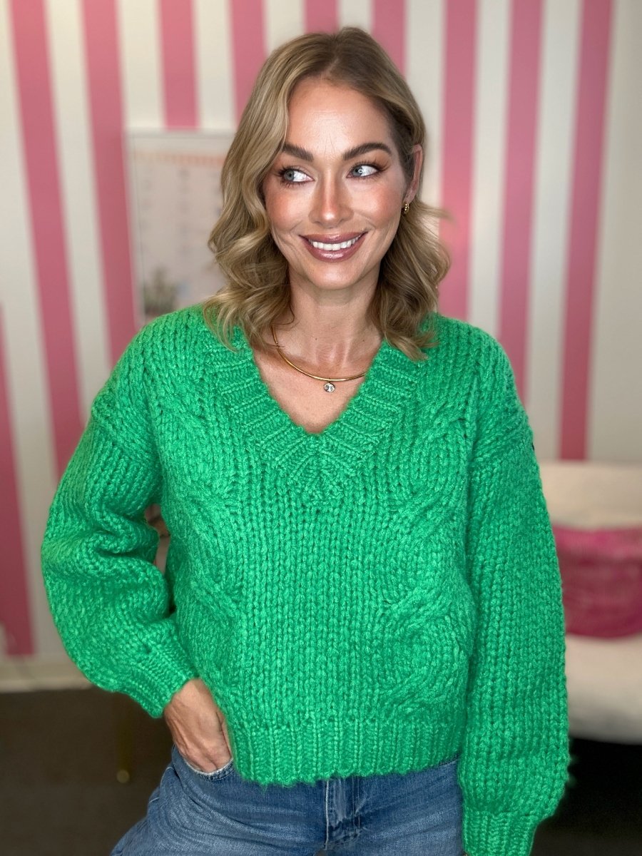 A woman wearing a bright green chunky cable-knit sweater and blue jeans, standing in front of a pink and white striped wall.