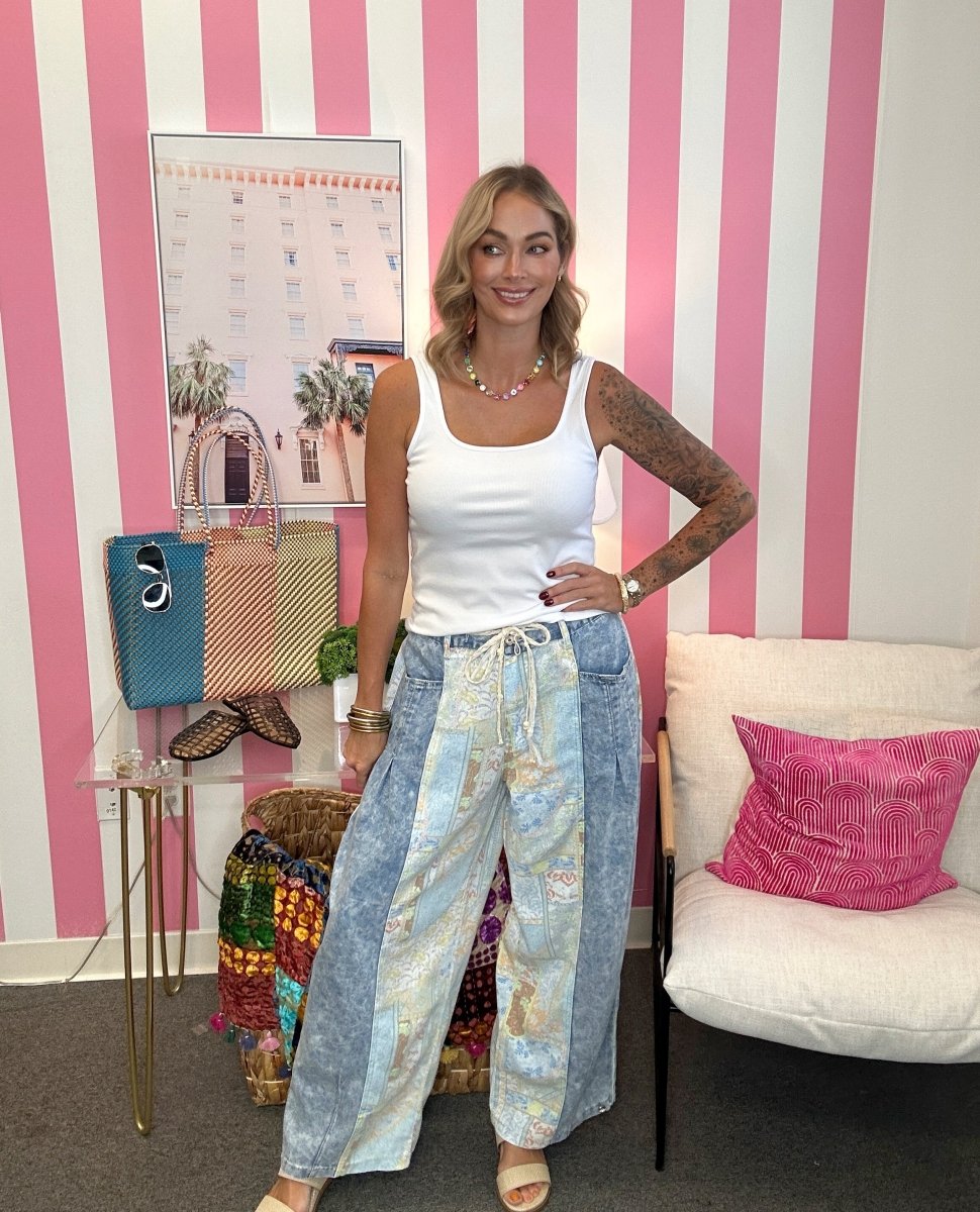 A woman stands in a room with pink and white striped walls, wearing a white tank top and wide-leg patchwork jeans with blue denim panels. She is posing next to a white armchair with a pink pillow and a small glass table holding colorful handbags.