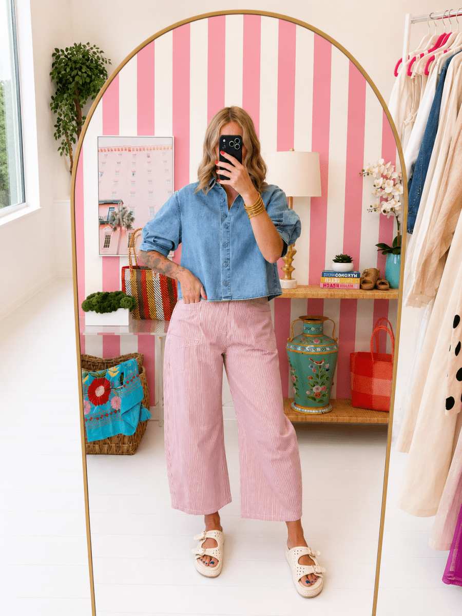 Person taking a mirror selfie wearing a blue shirt and pink pants in a room with striped walls and shelves.