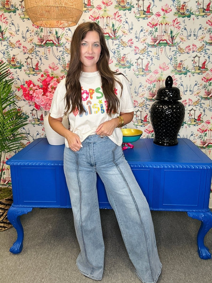 Woman wearing a colorful t-shirt and jeans standing in front of a patterned wall with a blue table.