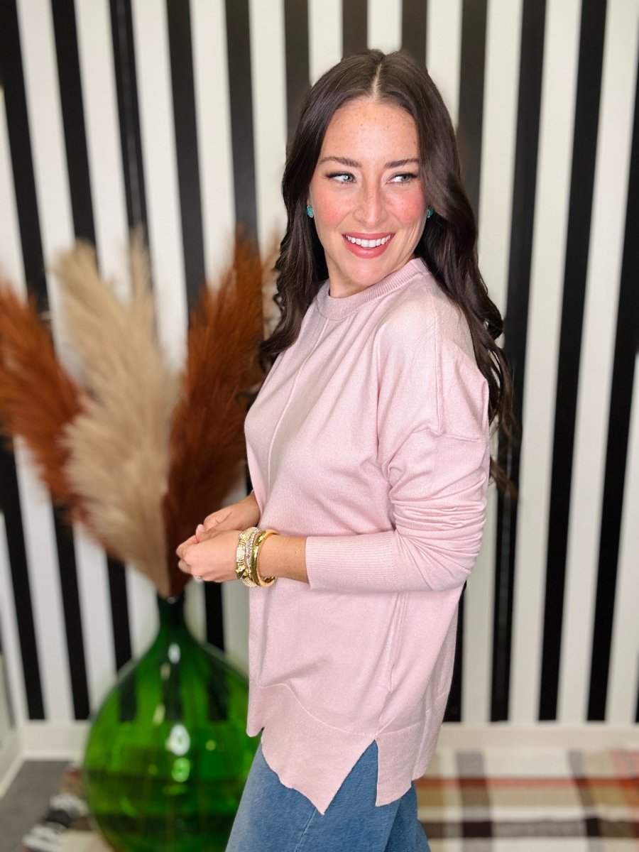 A woman wearing a light pink long-sleeve mock neck top and blue jeans, standing in front of a black and white striped wall with a green vase of pampas grass.