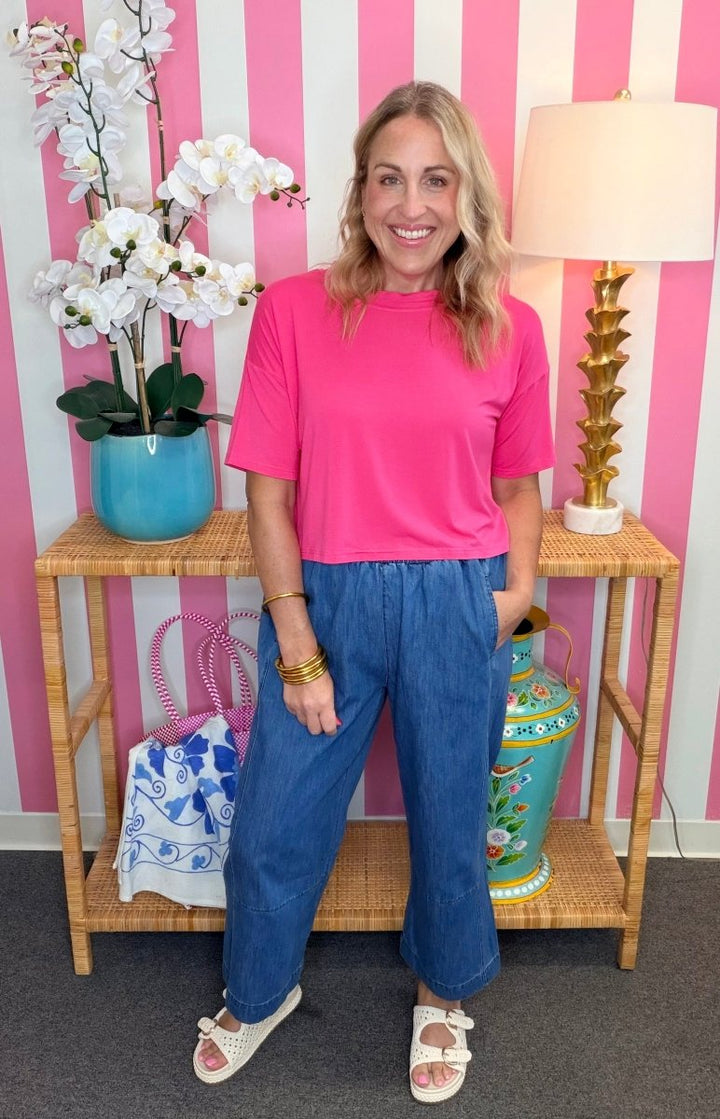 A woman stands indoors wearing a bright pink short-sleeve top and blue wide-leg pants, posed in front of a pink-and-white striped wall with a bamboo side table, lamp, and decorative vases.