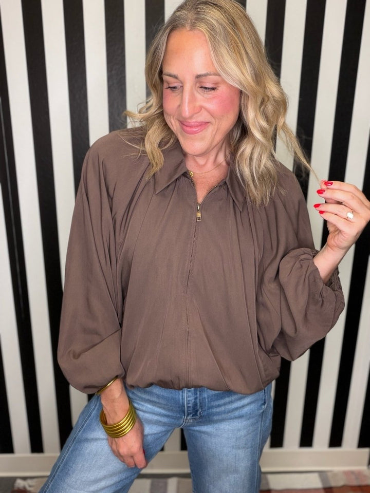 A woman wearing a brown long-sleeve blouse with a front zipper and blue jeans, standing in front of a black and white striped wall.