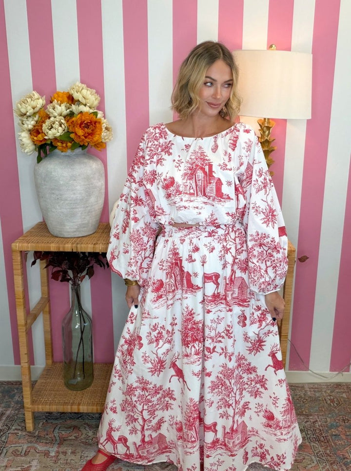 A woman wearing a long-sleeved white maxi dress with a red toile de Jouy print stands in front of a pink and white striped wall. She is posing next to a wooden side table with a vase of flowers and a lamp.