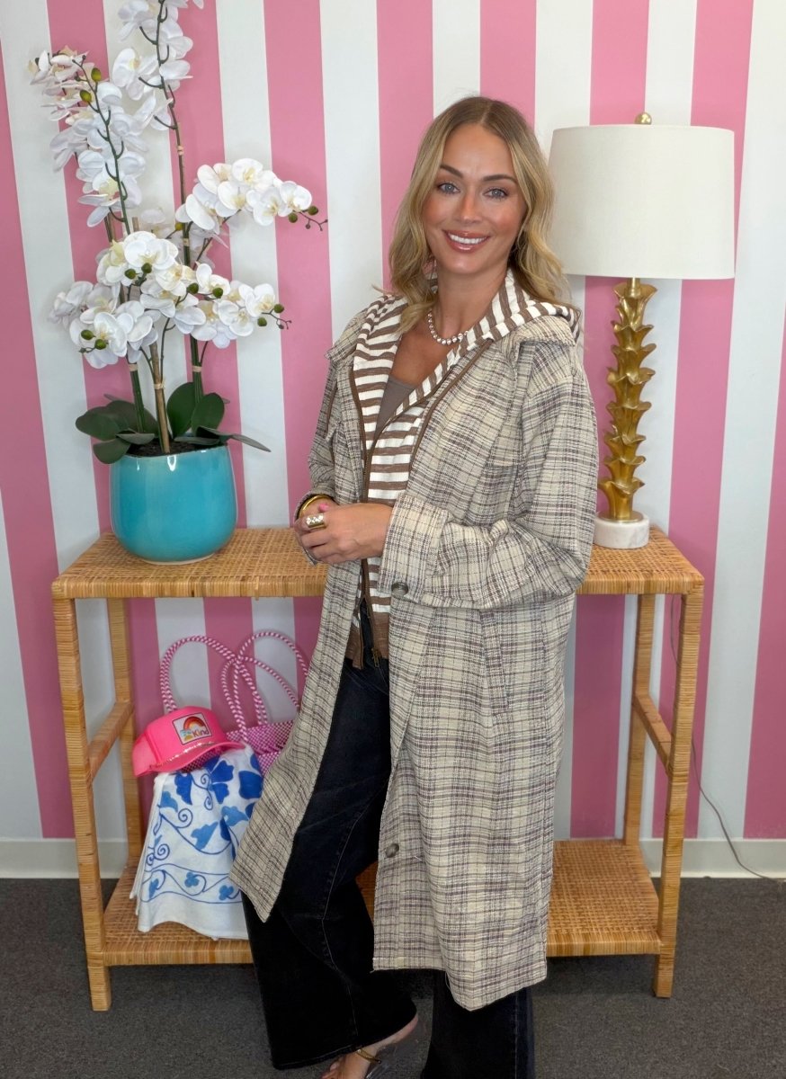 A woman stands indoors wearing a long beige plaid coat over a striped top and dark pants. She is posed in front of a pink-and-white striped wall with a wooden side table holding a turquoise orchid pot, a pink handbag, and a lamp.
