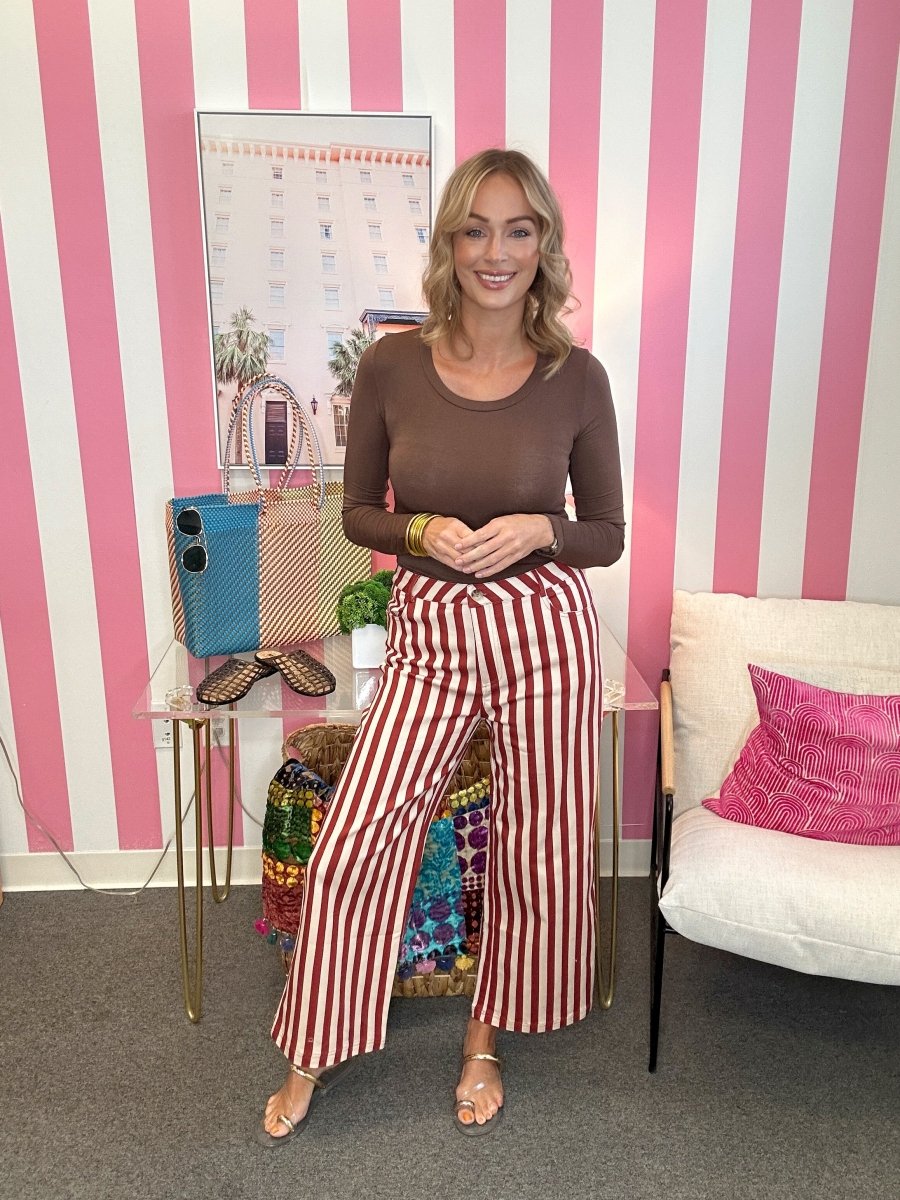 A woman stands in a room with pink and white striped wallpaper, wearing a brown long-sleeve top and red-and-white vertically striped wide-leg pants. She is posing next to a glass side table with a blue phone case and a white armchair with a pink pillow.