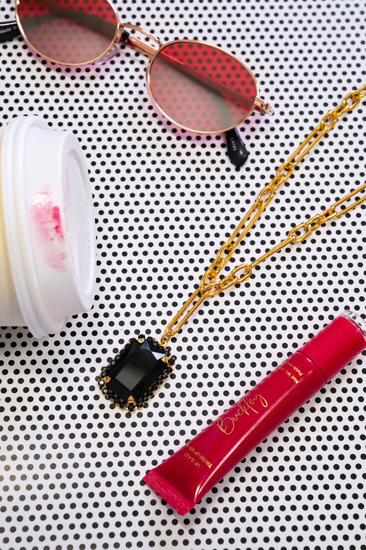 A gold chain necklace with a rectangular black gemstone pendant, styled on a black and white polka dot background alongside pink sunglasses, a pink lip product tube, and a white cosmetic container.