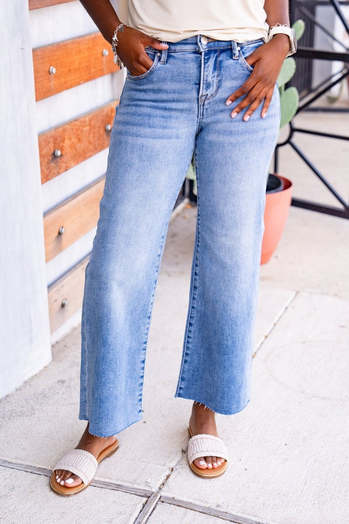 Person wearing light blue jeans standing on a concrete surface with a white wall and plant in the background.