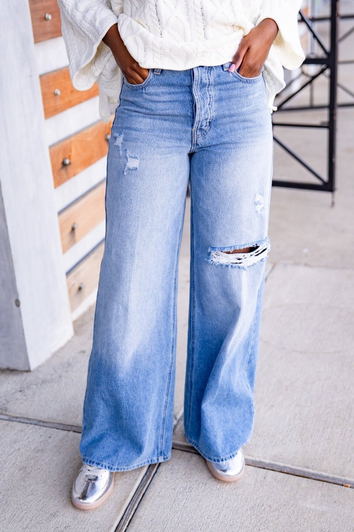 Person wearing light blue jeans with a distressed look, standing on a sidewalk.
