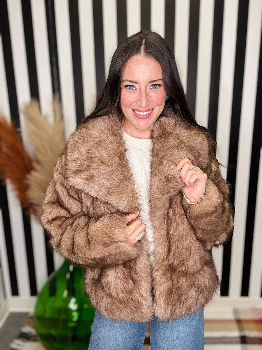 A woman smiling while wearing a brown faux fur jacket over a white top and blue jeans, standing in front of a black and white striped wall.