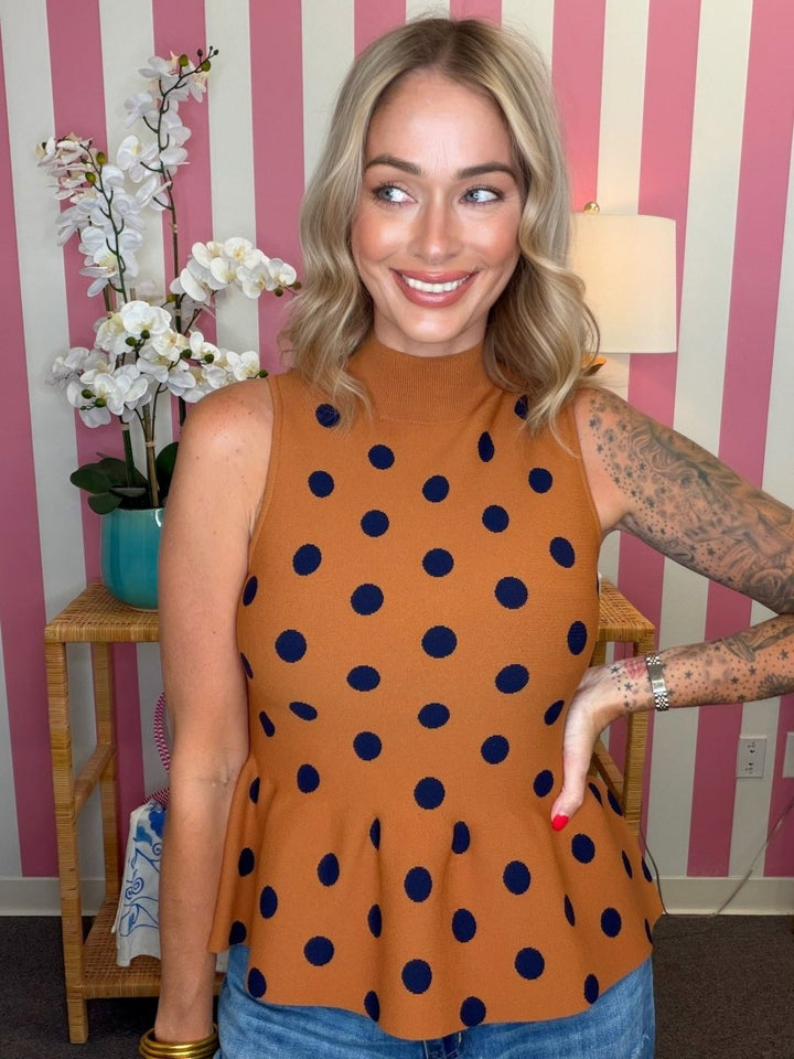 A woman wearing a sleeveless mock-neck top in rust orange with navy blue polka dots, standing in front of a pink and white striped wall.