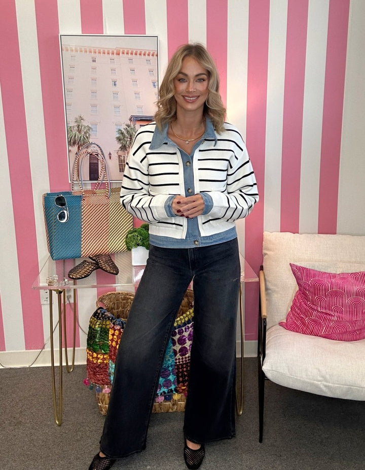 A woman stands in a pink and white striped room wearing a white cropped cardigan with black horizontal stripes and a light blue collar and button placket, paired with dark wide-leg jeans.