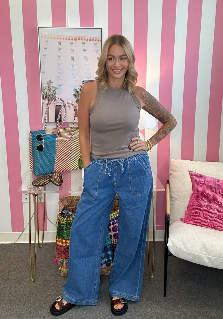 A woman stands in a room with pink and white vertical stripes, wearing a sleeveless taupe top and wide-leg blue jeans. She is smiling and posing, with a small table, colorful woven bag, and a white chair with a pink pillow in the background.