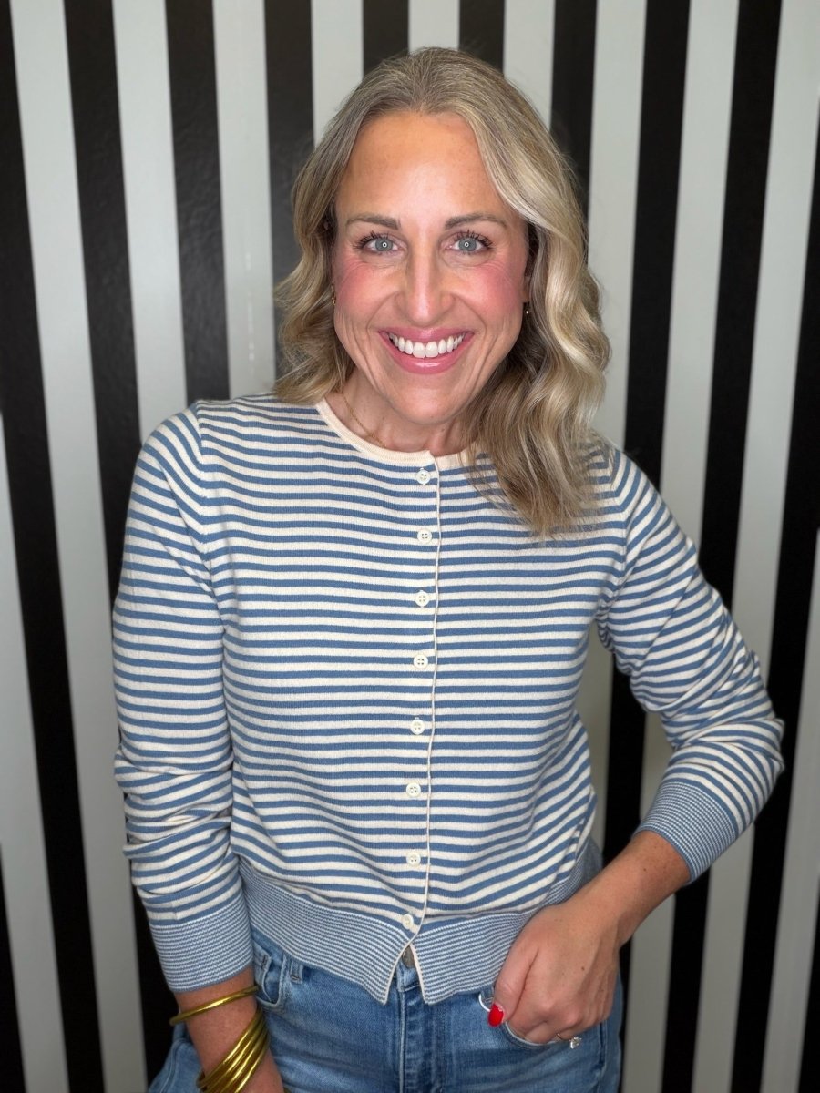 A smiling blonde woman wearing a blue and cream striped button-front cardigan over a light top and blue jeans, standing in front of a black and white vertically striped wall.