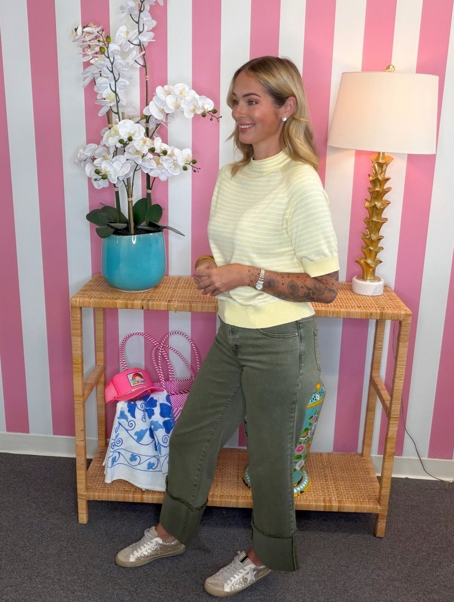 A woman modeling a pale yellow and white striped mock neck top with olive green cuffed pants and metallic silver sneakers, standing in front of a pink and white striped wall next to a bamboo console table with a lamp and a potted orchid.