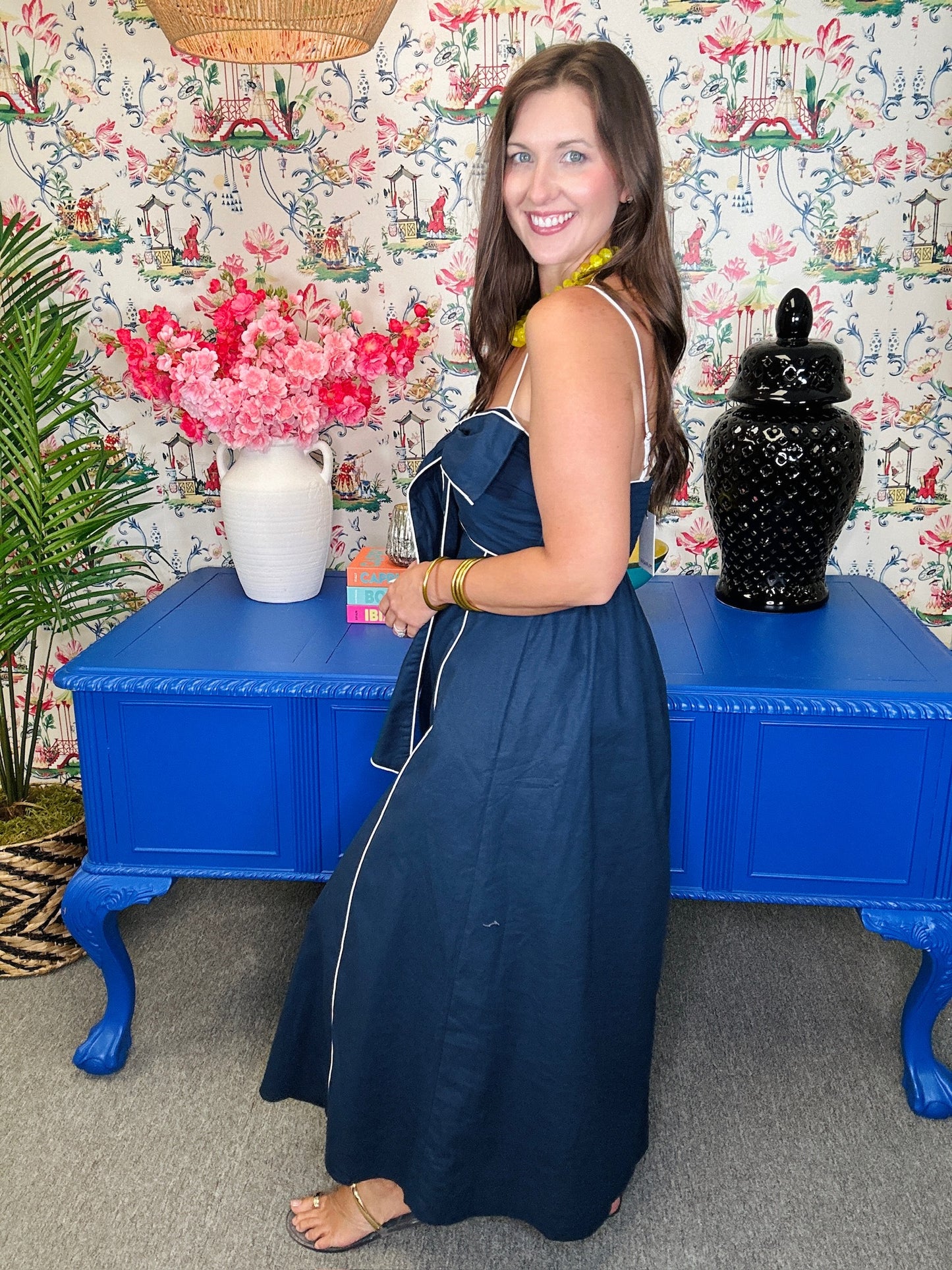 Woman in a blue dress standing in front of a colorful wall with floral patterns, a blue table, and decorative items.