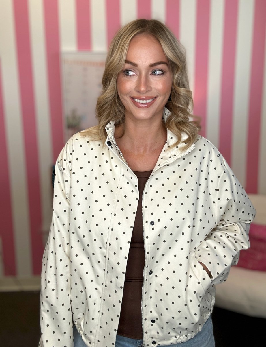 A woman with blonde hair wearing a cream-colored long-sleeve button-up shirt with black polka dots, paired with light blue jeans. She is standing in front of a pink and white striped wall.