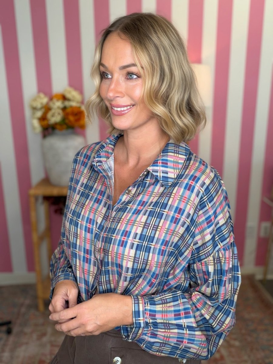 A woman with blonde wavy hair wearing a blue, pink, and white plaid button-down shirt, standing in front of a pink and white striped wall.